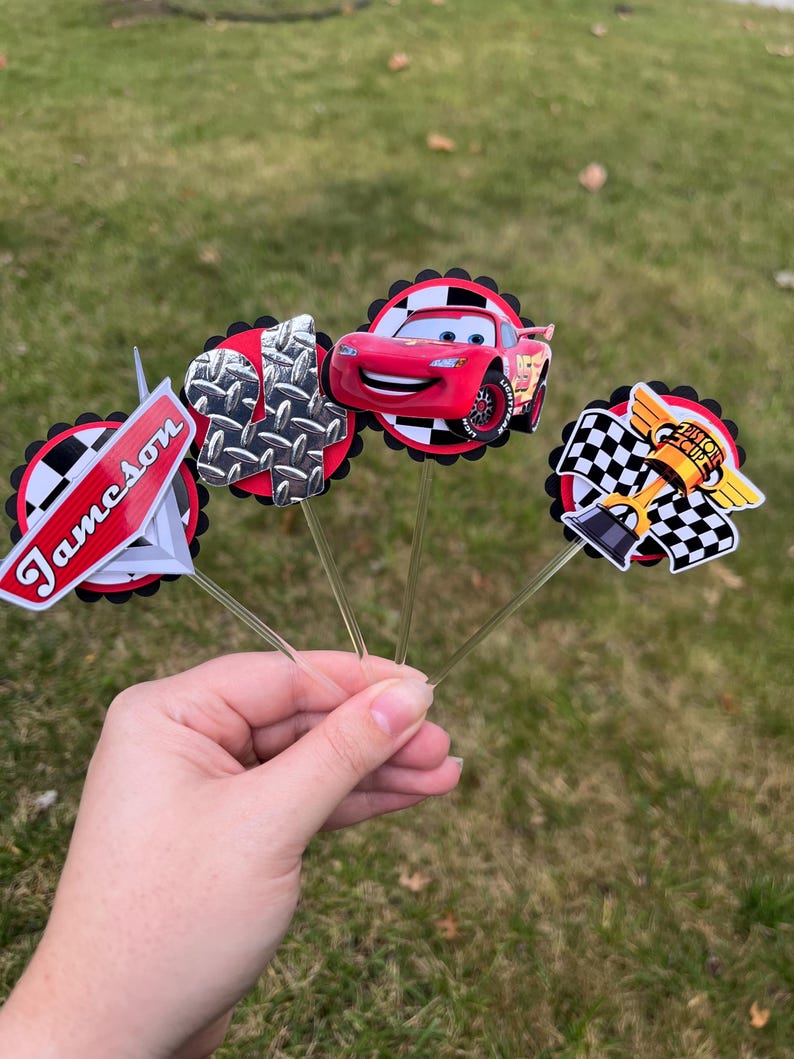 May include: A hand holds several decorative toppers featuring a red race car, a checkered flag, and a metal plate design. The toppers are on clear sticks, with a red and black circular border. One topper has the name "Jameson" on it.