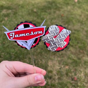 May include: Two cupcake toppers with a car theme. One has the name "Jameson" in red, the other has a silver diamond plate design. Both are on clear sticks, with red and black accents.