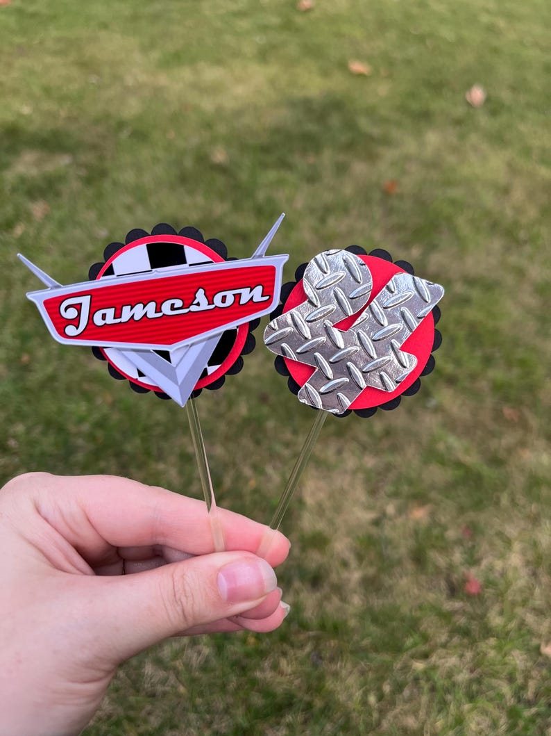 May include: Two cupcake toppers with a car theme. One has the name "Jameson" in red, the other has a silver diamond plate design. Both are on clear sticks, with red and black accents.