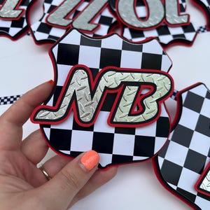 Puede incluir: Un recorte decorativo en forma de escudo con un patr&oacute;n de cuadros blancos y negros y un borde rojo. Las letras NB se muestran en una fuente plateada con textura de placa de diamante superpuesta. Otros recortes similares son visibles.