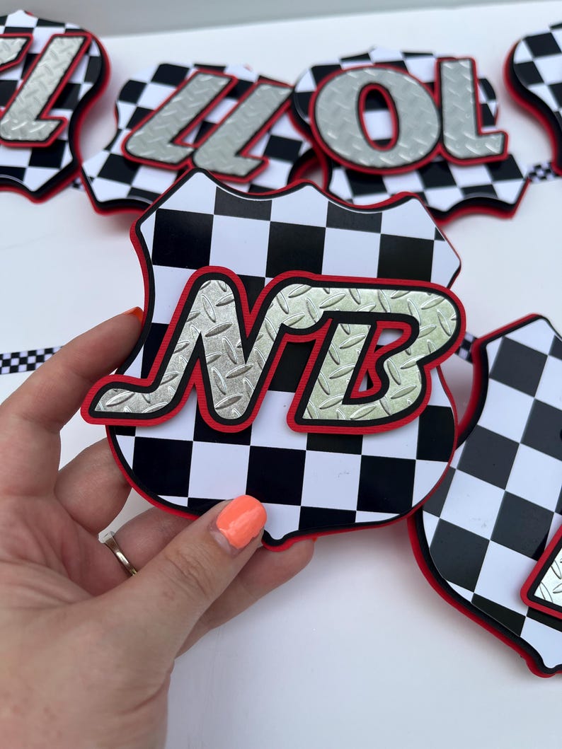 Puede incluir: Un recorte decorativo en forma de escudo con un patr&oacute;n de cuadros blancos y negros y un borde rojo. Las letras NB se muestran en una fuente plateada con textura de placa de diamante superpuesta. Otros recortes similares son visibles.