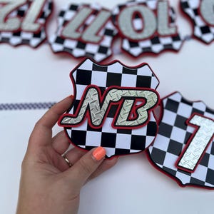 Puede incluir: Una pieza decorativa de pancarta con forma de escudo, con un patr&oacute;n de cuadros blancos y negros y un borde rojo. El centro presenta las letras NB en un estilo de placa de diamante plateada y texturizada. Otras piezas son visibles al fondo.