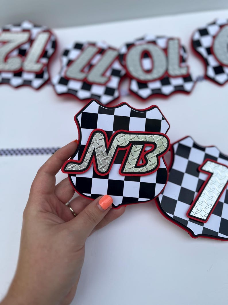 Puede incluir: Una pieza decorativa de pancarta con forma de escudo, con un patr&oacute;n de cuadros blancos y negros y un borde rojo. El centro presenta las letras NB en un estilo de placa de diamante plateada y texturizada. Otras piezas son visibles al fondo.