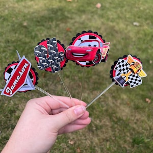 May include: A collection of four decorative cupcake toppers featuring designs from the animated movie Cars. The toppers include a red race car, a checkered flag, and the name "Jameson". Each topper is attached to a clear plastic stick.