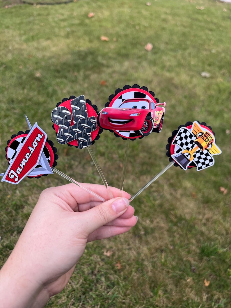 May include: A collection of four decorative cupcake toppers featuring designs from the animated movie Cars. The toppers include a red race car, a checkered flag, and the name "Jameson". Each topper is attached to a clear plastic stick.