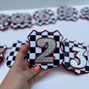 Puede incluir: Un recorte en forma de escudo con un patr&oacute;n de cuadros blancos y negros, un borde rojo y un n&uacute;mero dos plateado con textura de chapa estriada en el centro. El art&iacute;culo es parte de una serie de recortes numerados dispuestos en fila.