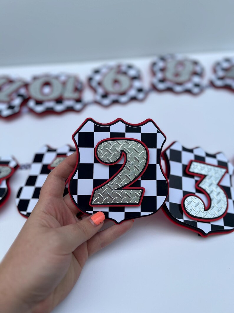 Puede incluir: Un recorte en forma de escudo con un patr&oacute;n de cuadros blancos y negros, un borde rojo y un n&uacute;mero dos plateado con textura de chapa estriada en el centro. El art&iacute;culo es parte de una serie de recortes numerados dispuestos en fila.