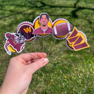 May include: Five cupcake toppers featuring a football player, a football, a helmet with the letter "W", and two different versions of the University of Washington logo. The toppers are on clear plastic sticks.