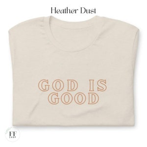 Op de afbeelding: Een beige T-shirt met de tekst "GOD IS GOOD" in bruin gedrukt op de voorkant.