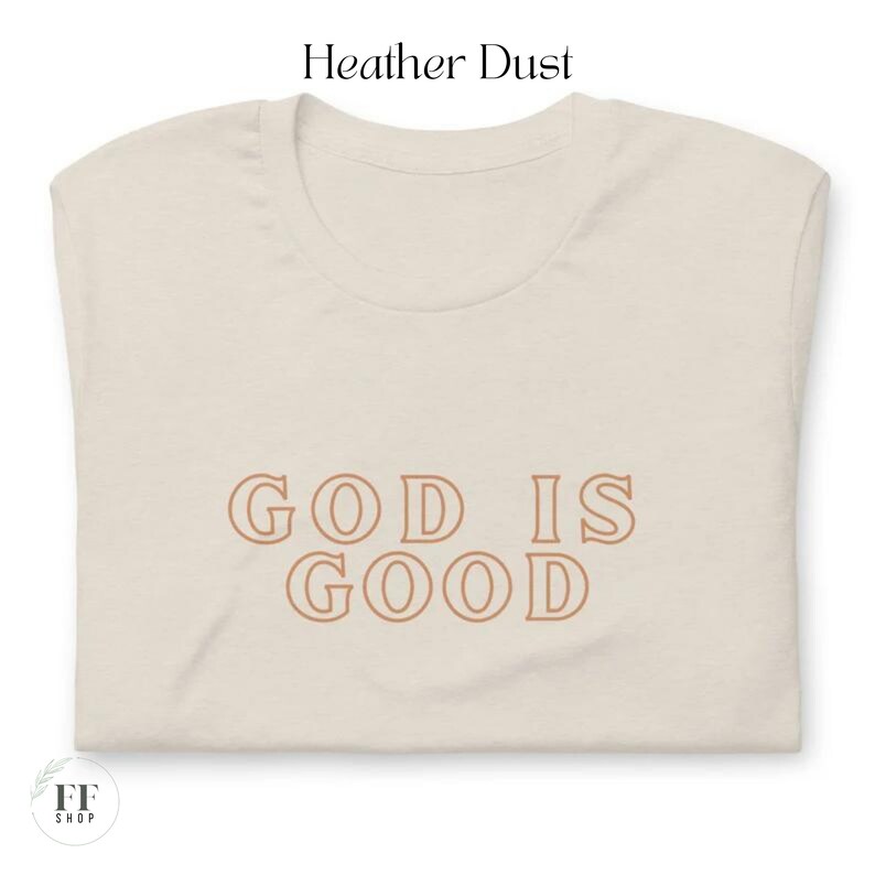 Op de afbeelding: Een beige T-shirt met de tekst "GOD IS GOOD" in bruin gedrukt op de voorkant.