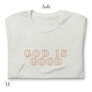 Op de afbeelding: Een lichtgrijs T-shirt met de tekst "GOD IS GOOD" in bruin gedrukt op de voorkant.