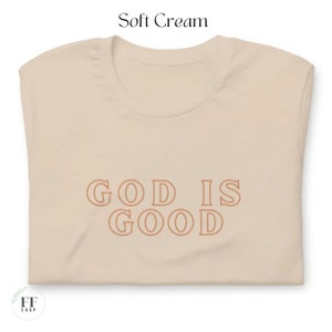 Op de afbeelding: Een zachtcr&eacute;mekleurig T-shirt met de tekst "GOD IS GOOD" in bruin gedrukt.