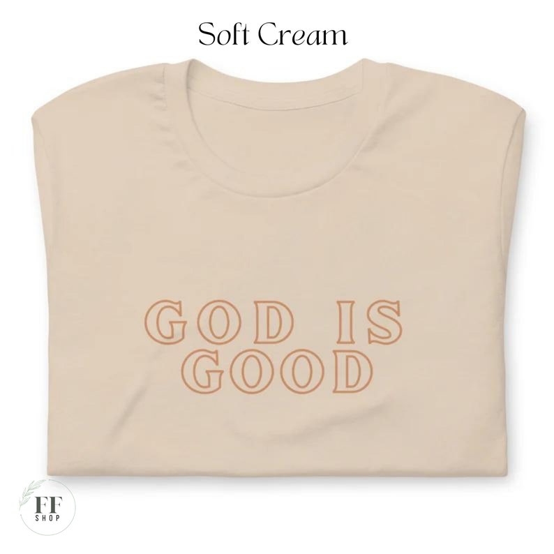 Op de afbeelding: Een zachtcr&eacute;mekleurig T-shirt met de tekst "GOD IS GOOD" in bruin gedrukt.
