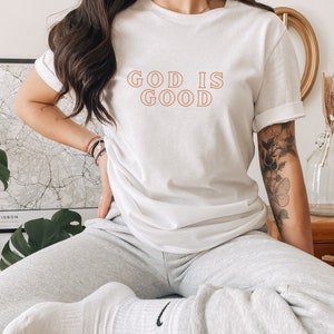 Op de afbeelding: Wit T-shirt met de tekst "GOD IS GOOD" in oranje omrande letters.