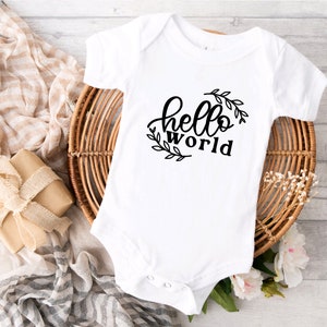 Peut inclure: Body bébé blanc avec l'inscription "hello world" en noir, entourée d'une couronne de feuilles. Le body est présenté sur un panier en osier, avec un cadeau emballé et du tissu en arrière-plan.