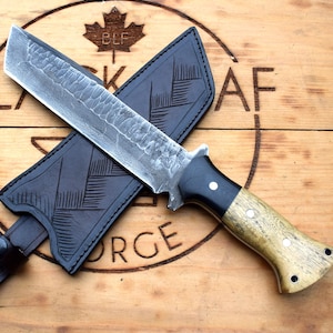 Puede incluir: Un cuchillo grande, forjado a mano, con una hoja texturizada y un mango de madera, sobre una superficie de madera. El cuchillo va acompañado de una funda de cuero negro. Las palabras "BLACK LEAF FORGE" son visibles.
