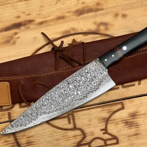 Puede incluir: Un cuchillo de chef de acero Damasco con mango negro y rojo, en una funda de cuero marrón. El cuchillo tiene un patrón ondulado y es muy afilado.