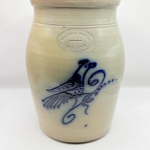 May include: A beige stoneware crock with a smooth finish and a brown rim. The front features a blue cobalt slip-trailed design of a bird perched on a decorative vine. An oval stamp reads Beaumont Pottery York, Maine in the upper center.