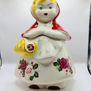 May include: A ceramic cookie jar shaped like a figure wearing a red hood and cape. The figure has yellow hair, a white apron with gold star accents, and a yellow basket. The skirt features red and white rose patterns on a cream-coloured base.