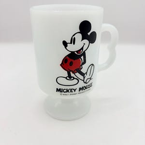 May include: A white milk glass pedestal mug featuring a graphic of Mickey Mouse wearing red shorts. The text MICKEY MOUSE and &copy; WALT DISNEY PRODUCTIONS is printed in black below the character. The mug has a curved handle and a circular base.