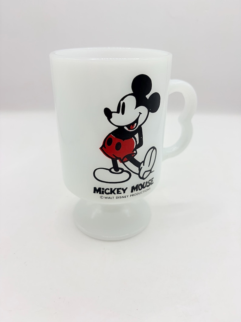 May include: A white milk glass pedestal mug featuring a graphic of Mickey Mouse wearing red shorts. The text MICKEY MOUSE and &copy; WALT DISNEY PRODUCTIONS is printed in black below the character. The mug has a curved handle and a circular base.