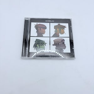 May include: A compact disc case featuring the album "Demon Days" by Gorillaz. The cover art displays four illustrated character portraits in a grid, with the band's name at the top. The disc is encased in clear plastic.