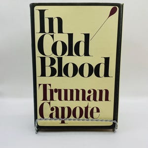 May include: A hardback book titled "In Cold Blood" by Truman Capote. The book has a light yellow cover with black and burgundy lettering. The book is held in a silver metal book stand.