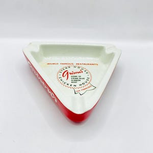 May include: Triangular ashtray with a red border and white interior. The ashtray features the text "World Famous Restaurants" and "Grison's Steak House" with additional text and a logo. The ashtray is a vintage item.