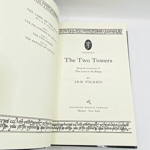 May include: Open book with the title "The Two Towers" by J.R.R. Tolkien. The book's pages are cream-colored with black text and decorative borders. The cover is black. The book is part of "The Lord of the Rings" series.