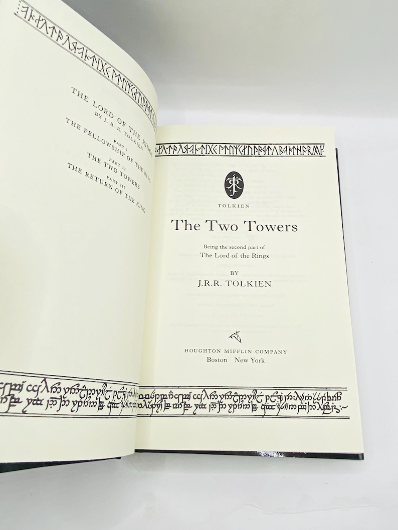 May include: Open book with the title "The Two Towers" by J.R.R. Tolkien. The book's pages are cream-colored with black text and decorative borders. The cover is black. The book is part of "The Lord of the Rings" series.