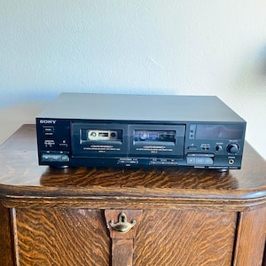 Könnte beinhalten: Schwarzes Sony Stereo-Kassettendeck mit zwei Kassettenfächern, verschiedenen Knöpfen und einem Lautstärkeregler. Eine Kassette ist in einem Fach eingelegt. Das Deck steht auf einem Holzschrank.