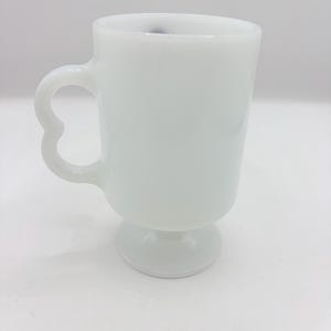 May include: A solid white milk glass mug featuring a smooth, cylindrical body, a pedestal base, and a distinct heart-shaped handle. The mug has a glossy finish and a clean, simple design with no additional patterns, colors, or text visible on the surface.
