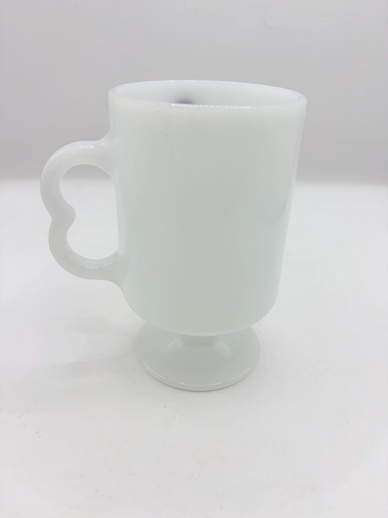 May include: A solid white milk glass mug featuring a smooth, cylindrical body, a pedestal base, and a distinct heart-shaped handle. The mug has a glossy finish and a clean, simple design with no additional patterns, colors, or text visible on the surface.