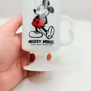 May include: A white milk glass pedestal mug featuring a graphic of Mickey Mouse in red shorts. The text MICKEY MOUSE is printed in black below the character, followed by the smaller text &copy;WALT DISNEY PRODUCTIONS. The mug has a heart-shaped handle.