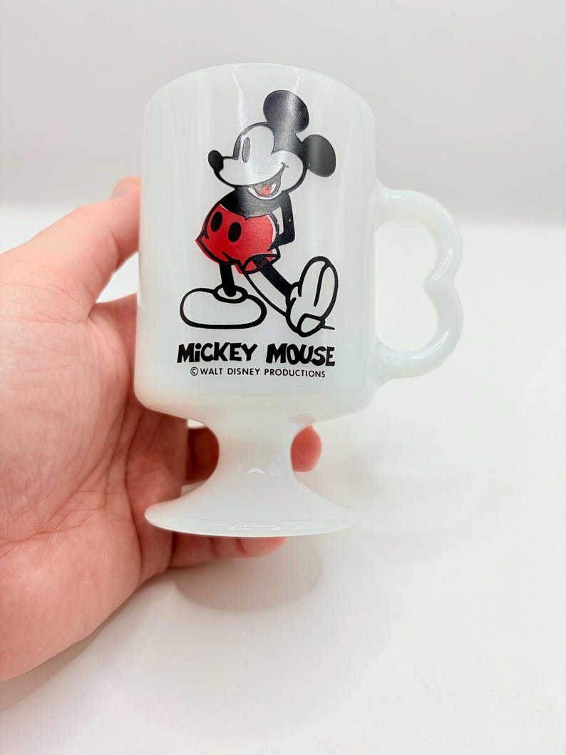 May include: A white milk glass pedestal mug featuring a graphic of Mickey Mouse in red shorts. The text MICKEY MOUSE is printed in black below the character, followed by the smaller text &copy;WALT DISNEY PRODUCTIONS. The mug has a heart-shaped handle.