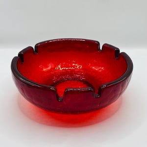 May include: A red glass ashtray with a textured surface and four indentations around the rim.