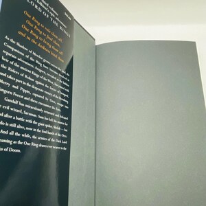 May include: An open book with the title "THE LORD OF THE RINGS" visible on the left page. The text includes the phrase "One Ring to rule them all." The book's pages are gray.