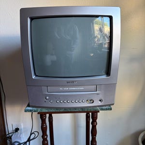 May include: A silver Toshiba TV/VCR combination unit featuring a cathode ray tube screen, a front-loading VCR slot, integrated speakers on the sides, and a control panel with circular buttons and input ports located below the tape compartment.