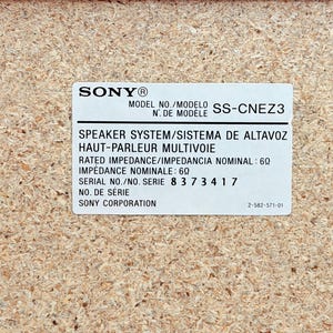 May include: A rectangular white product label on a light brown particle board surface. The label features the Sony logo, model number SS-CNEZ3, speaker system specifications, a rated impedance of 6 ohms, serial number 8 3 7 3 4 1 7, and Sony Corporation.