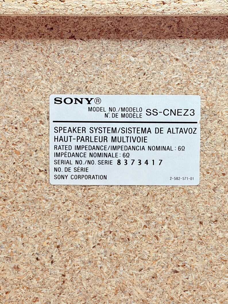 May include: A rectangular white product label on a light brown particle board surface. The label features the Sony logo, model number SS-CNEZ3, speaker system specifications, a rated impedance of 6 ohms, serial number 8 3 7 3 4 1 7, and Sony Corporation.