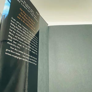 May include: A hardcover book titled "The Lord of the Rings" is shown, with text visible on the cover and pages. The cover is black with white text, and the pages are a light gray color. The book is slightly open, revealing the text on the pages.