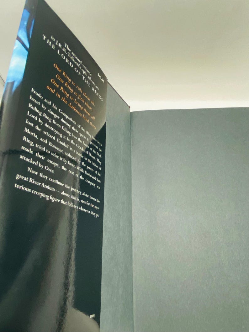 May include: A hardcover book titled "The Lord of the Rings" is shown, with text visible on the cover and pages. The cover is black with white text, and the pages are a light gray color. The book is slightly open, revealing the text on the pages.