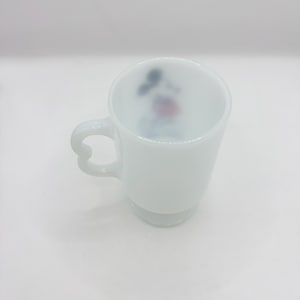 May include: A white milk glass mug featuring a heart-shaped handle and a small, colorful graphic of a cartoon character printed on the interior wall. The mug has a smooth, opaque finish and a pedestal base, standing upright against a plain background.