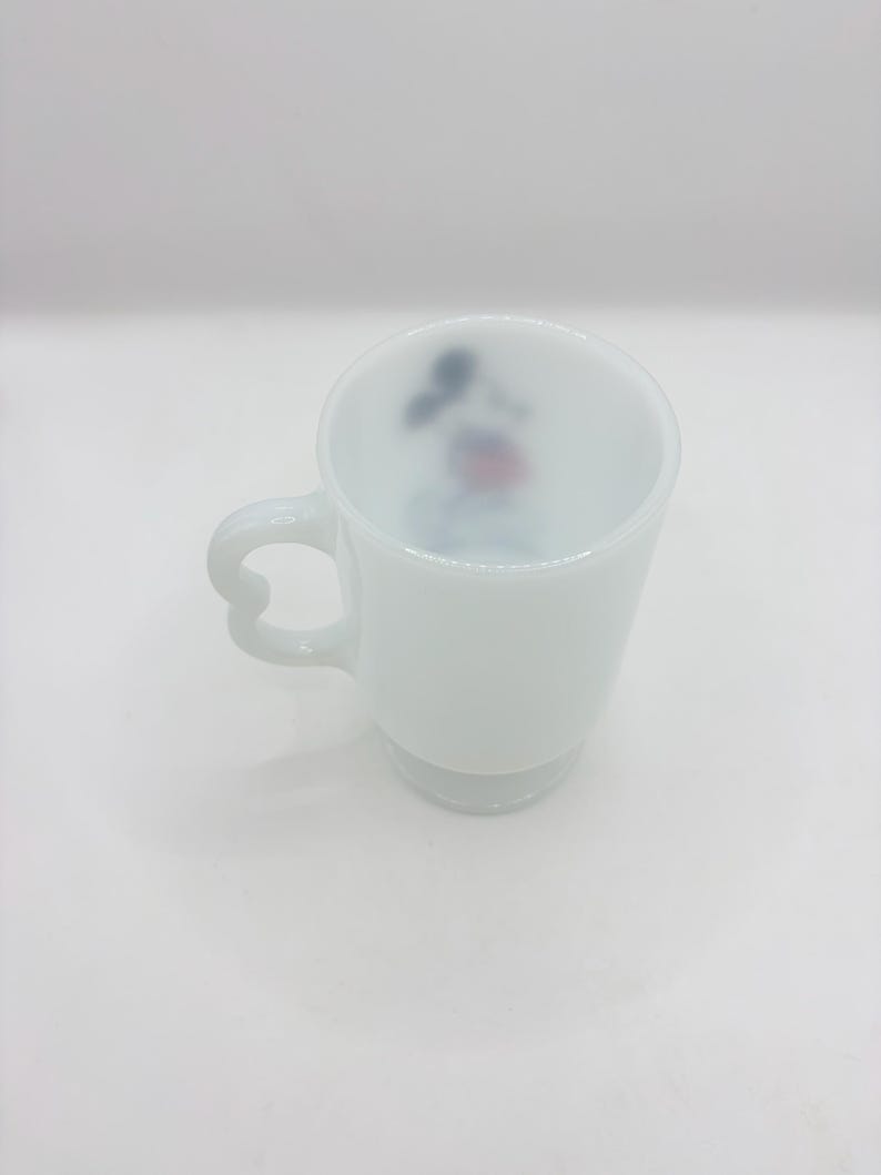 May include: A white milk glass mug featuring a heart-shaped handle and a small, colorful graphic of a cartoon character printed on the interior wall. The mug has a smooth, opaque finish and a pedestal base, standing upright against a plain background.