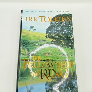 May include: A hardcover book titled "The Fellowship of the Ring" by J.R.R. Tolkien. The cover features a landscape with green hills, a lake, and the title in gold lettering. The book is part of "The Lord of the Rings" series.