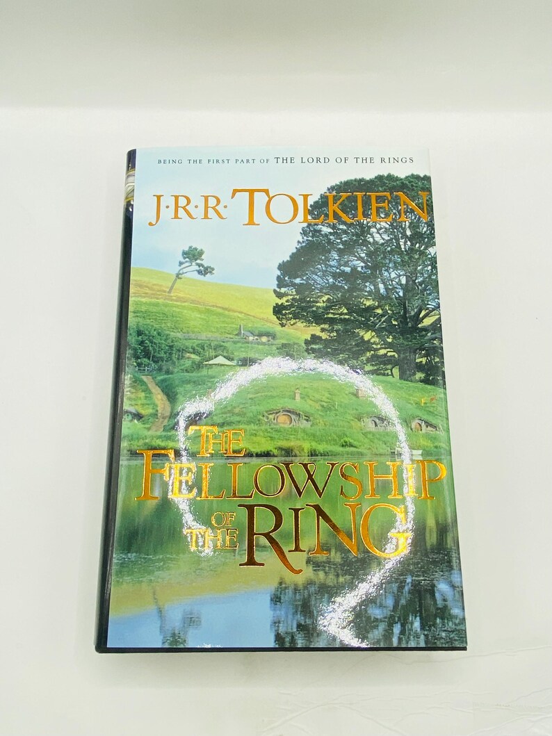 May include: A hardcover book titled "The Fellowship of the Ring" by J.R.R. Tolkien. The cover features a landscape with green hills, a lake, and the title in gold lettering. The book is part of "The Lord of the Rings" series.