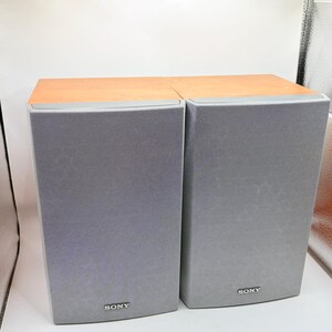 May include: A pair of rectangular Sony speakers featuring light wood-toned top panels and grey fabric mesh grilles covering the front. Each speaker displays the Sony brand name in small white lettering at the bottom centre of the front panel.