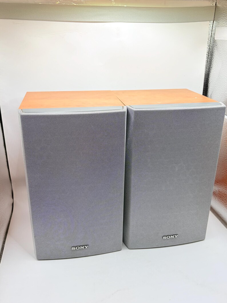 May include: A pair of rectangular Sony speakers featuring light wood-toned top panels and grey fabric mesh grilles covering the front. Each speaker displays the Sony brand name in small white lettering at the bottom centre of the front panel.