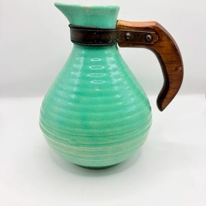 Peut inclure: Une carafe en céramique turquoise avec une texture horizontale nervurée. Le récipient présente une poignée en bois brun foncé fixée au col par une bande métallique sombre maintenue par deux vis visibles. La carafe a un corps large et arrondi.
