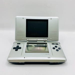 Può includere: Console di gioco portatile Nintendo DS argentata, aperta. Il dispositivo ha due schermi, un pad direzionale e pulsanti di azione. Il logo Nintendo DS è visibile sullo schermo inferiore.